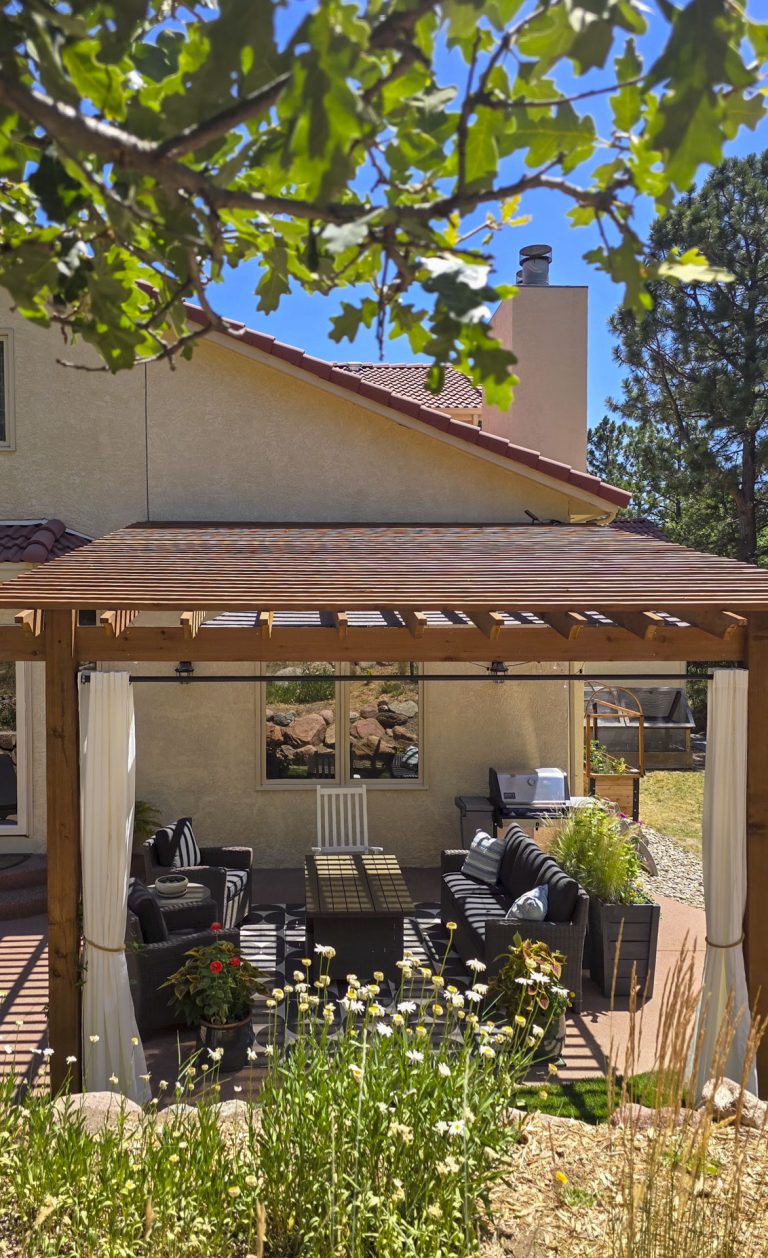 Wooden pergola with shade structure covering outdoor seating area, white flowers and greenery visible