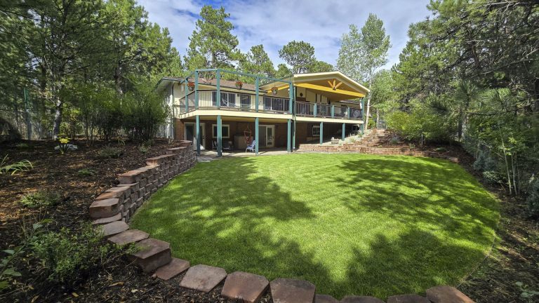 Modern home with wraparound deck and stone pathway surrounded by mature trees and manicured lawn