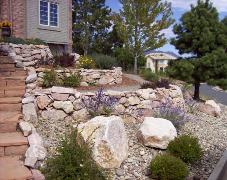 Residential rock garden with stacked stone wall, large boulders, and ornamental shrubs along brick pathway near house