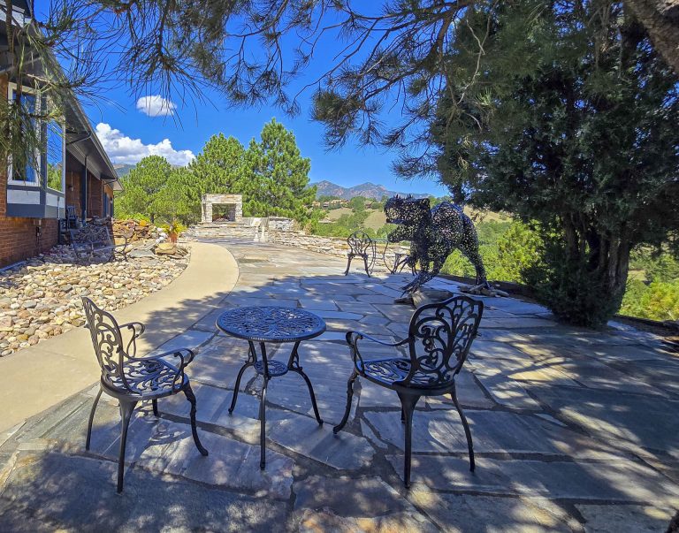 Black ornate outdoor dining table and chairs on a stone patio with trees and buildings in the background