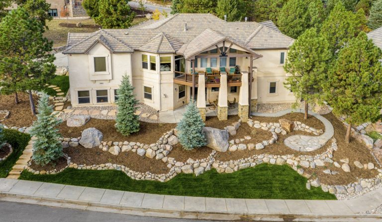 Aerial view of a large two-story stone mansion with columns, manicured landscaping, and curved driveway surrounded by trees
