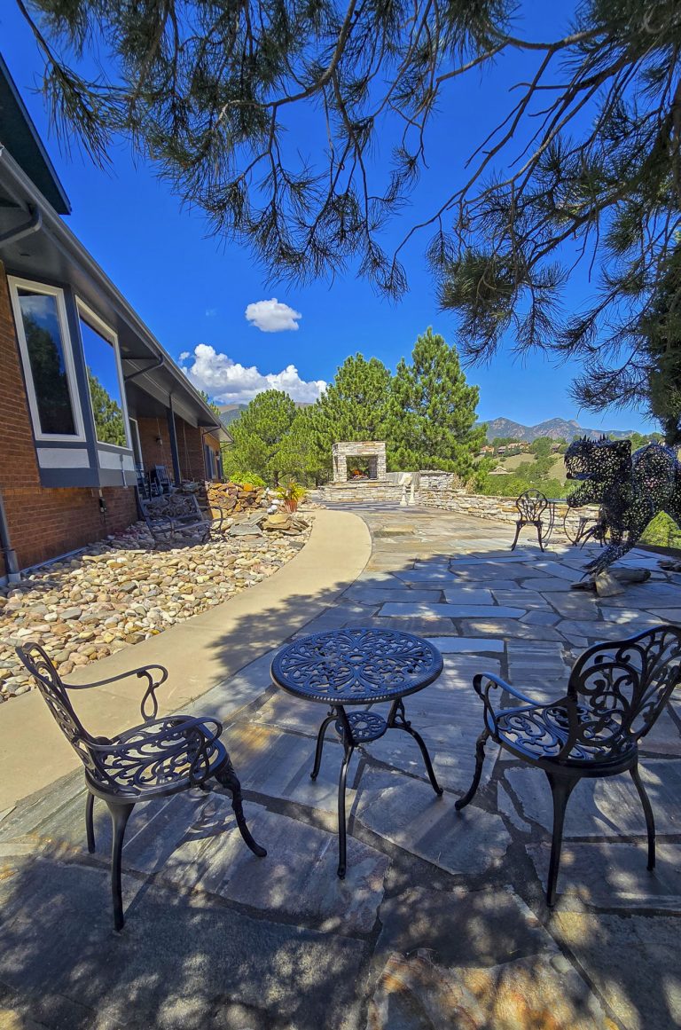 Outdoor patio area with metal chairs casting shadows on gravel, brick house, and trees under blue sky