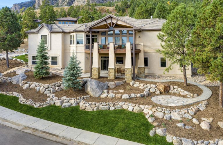 Modern two-story residential home with stone columns at entrance, surrounded by landscaped grounds with trees and rocks