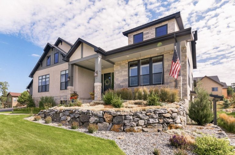 Two-story residential home with stone foundation, manicured lawn, landscaping, and American flag on a sunny day
