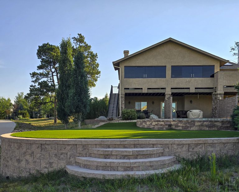 Two-story house with covered porch overlooking manicured lawn and circular stone patio surrounded by trees
