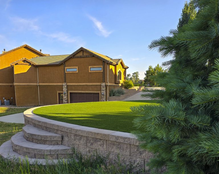 Brown barn with yellow trim, curved stone retaining wall, manicured lawn, and evergreen trees under blue sky