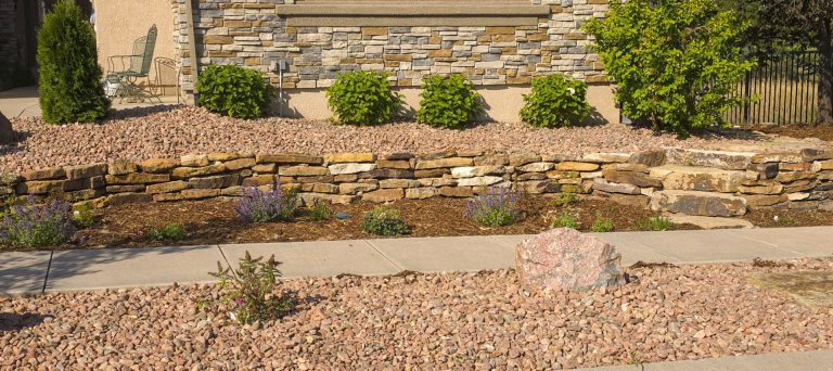 Curved stone retaining wall with gravel ground and green shrubs along landscaped garden bed