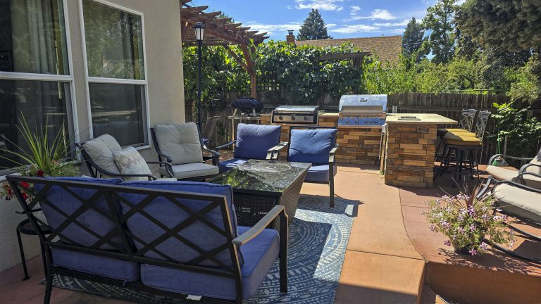 Modern outdoor patio with blue furniture, fireplace, and lush green landscaping under blue sky