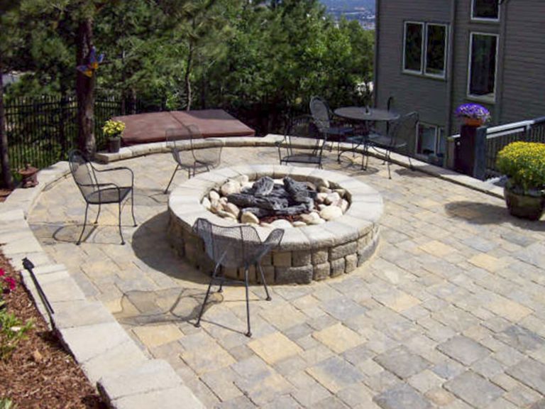 Circular stone patio with built-in fire pit, metal chairs, and lounge area surrounded by landscaping
