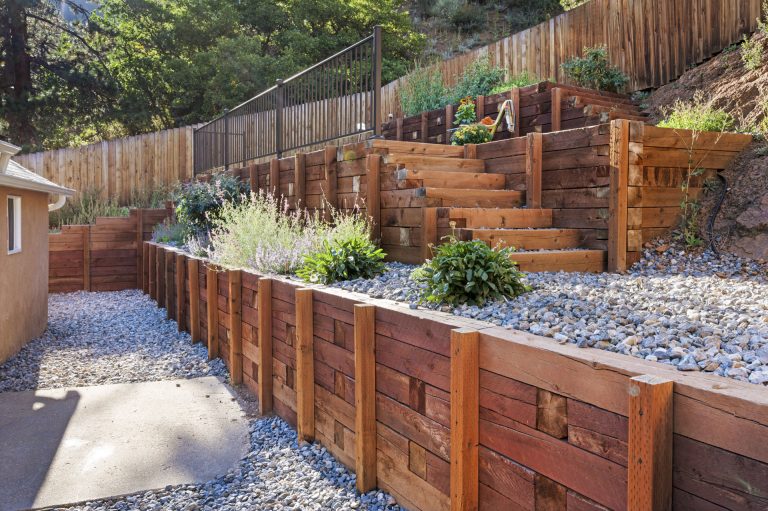 Wooden raised garden beds with gravel pathways, plants, and tiered wooden fence on hillside landscape