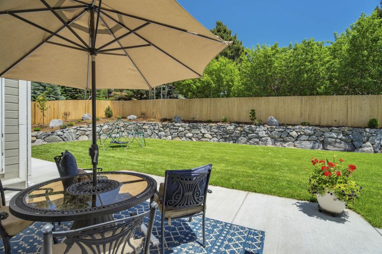 Backyard patio with outdoor dining set under beige umbrella, concrete patio, manicured lawn, and wooden fence with