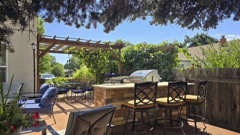 Backyard patio with wooden dining table, chairs, grill, and shade from trees under blue sky