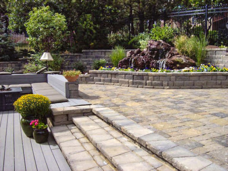 Backyard patio with stone pavers, steps, raised garden beds, potted plants, and wooden deck surrounded by landscaping