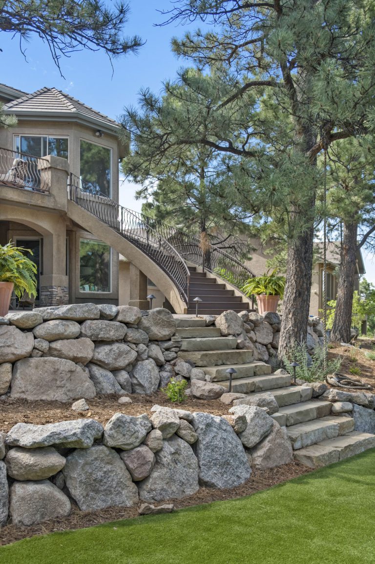 Stone steps and boulders lead up to a large residential home surrounded by tall pine trees on a manicured lawn