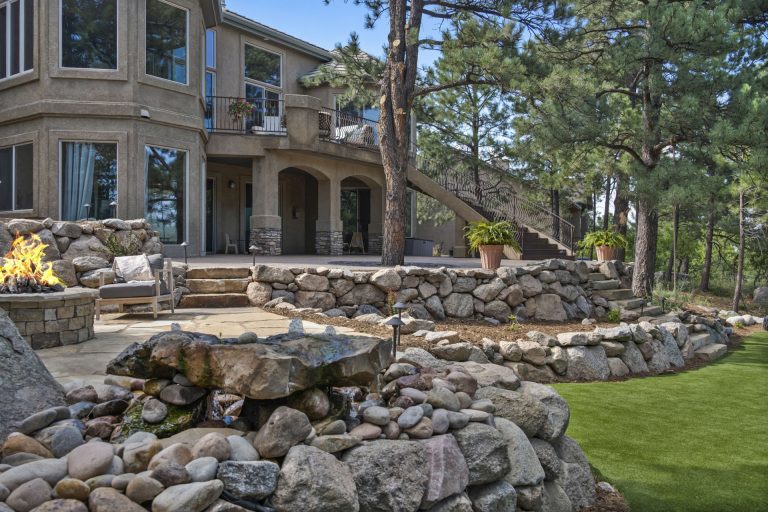 Luxury home exterior with stone landscaping, manicured lawn, and tall pine trees in background