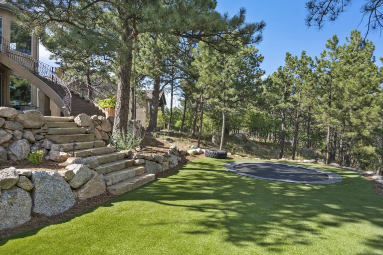 Spacious backyard with manicured green lawn, stone retaining wall, wooden deck, and tall pine trees under clear blue sky
