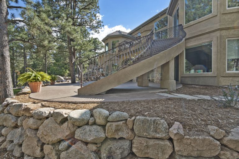 Modern home exterior with stone patio, wooden deck, large windows, and surrounding pine trees on a clear day