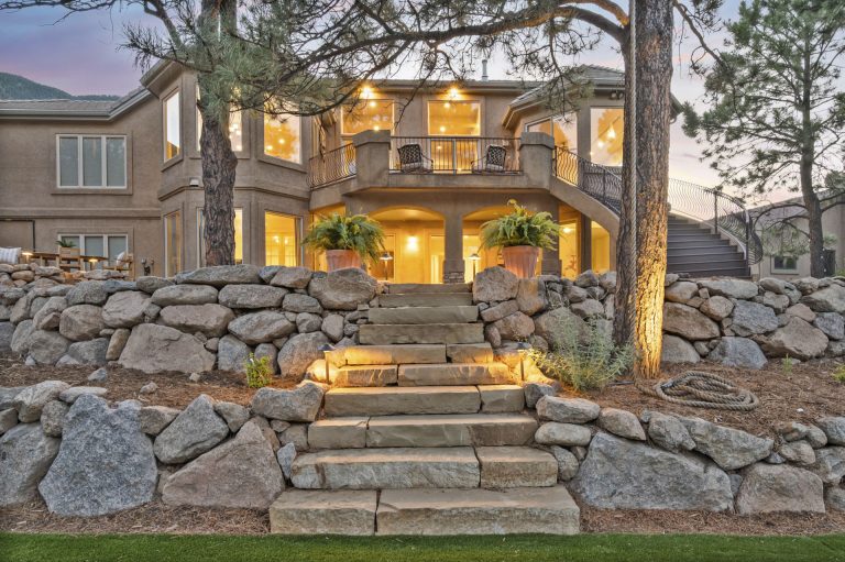 Stone house with illuminated windows and entrance steps at dusk surrounded by landscaping rocks