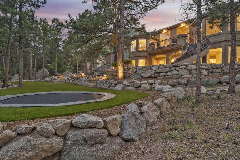 Modern stone house with illuminated windows at dusk, featuring a round pool and manicured lawn surrounded by tall pine trees