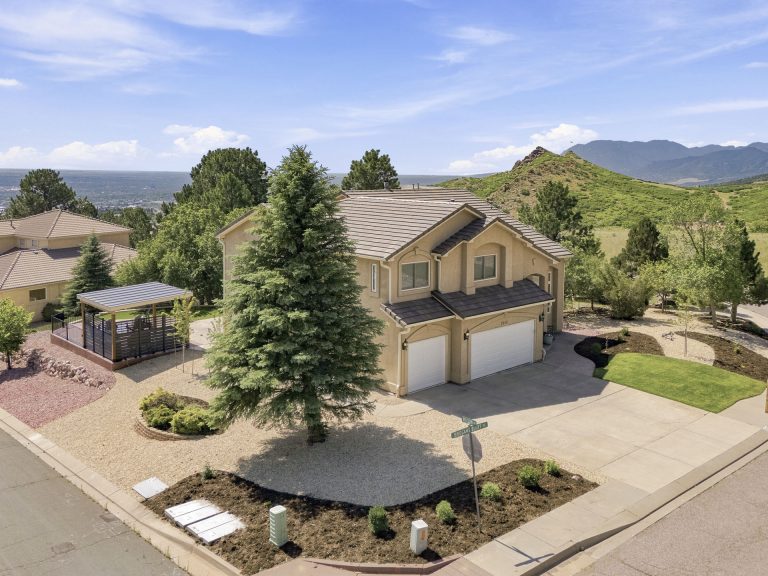 Aerial view of two-story house with attached garage, circular driveway, and landscaped yard in rural setting with mountains