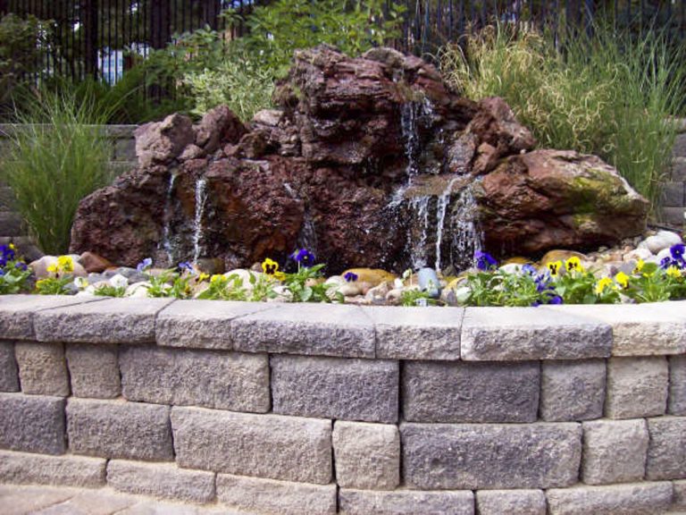 Decorative rock water fountain feature with lava rock in landscaped garden bed surrounded by stone retaining wall