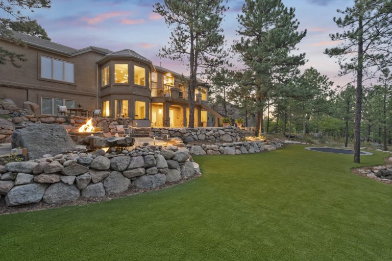 Modern home with illuminated windows at dusk, featuring stone foundation, manicured lawn, and evergreen trees