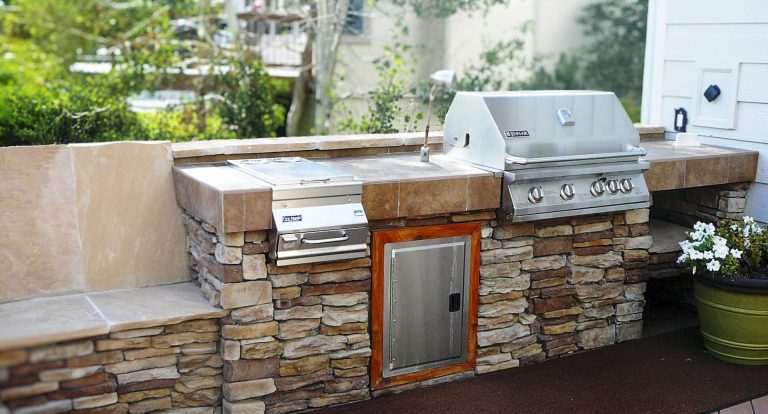 Outdoor kitchen with stone countertop, built-in grill, storage cabinet, and white flowers in a backyard setting