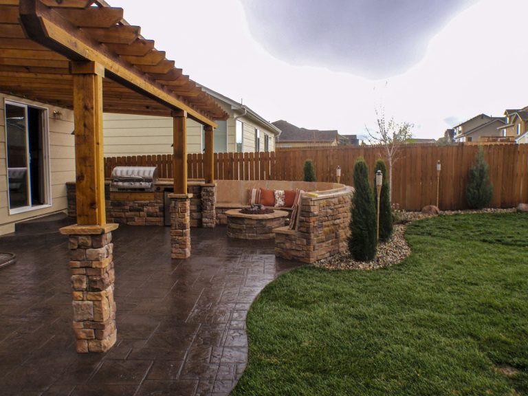 Backyard patio with wooden pergola, stone columns, dark pavers, and manicured lawn under cloudy sky